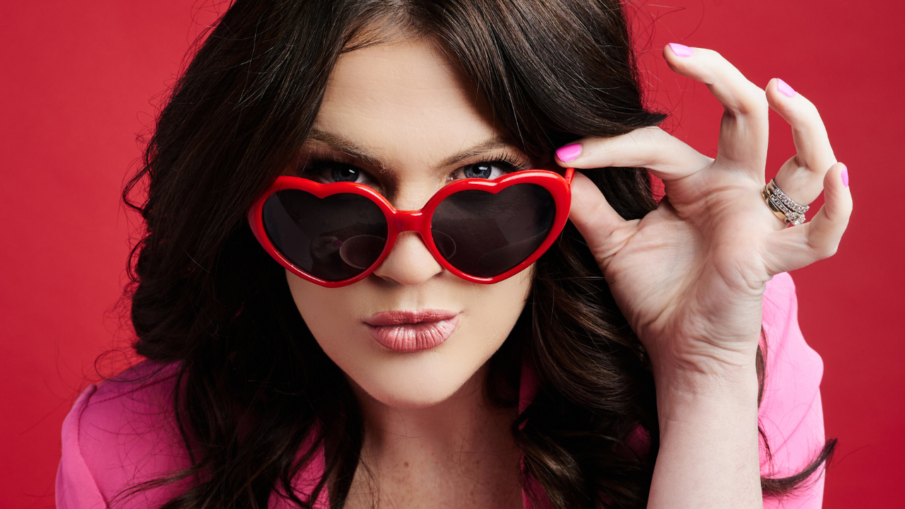 Emily standing in front of a red background, wearing a pink blazer and red heart shaped sunglasses. She is holding the sunglasses with one hand and a lip pattern mug in the other.