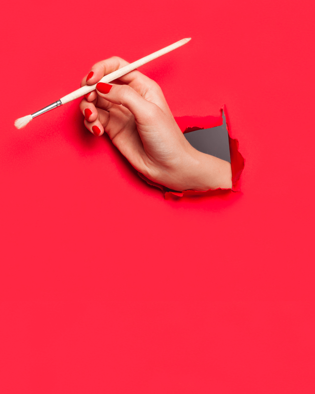 Red backdrop with hole for a hand with red nail polish holding a paintbrush