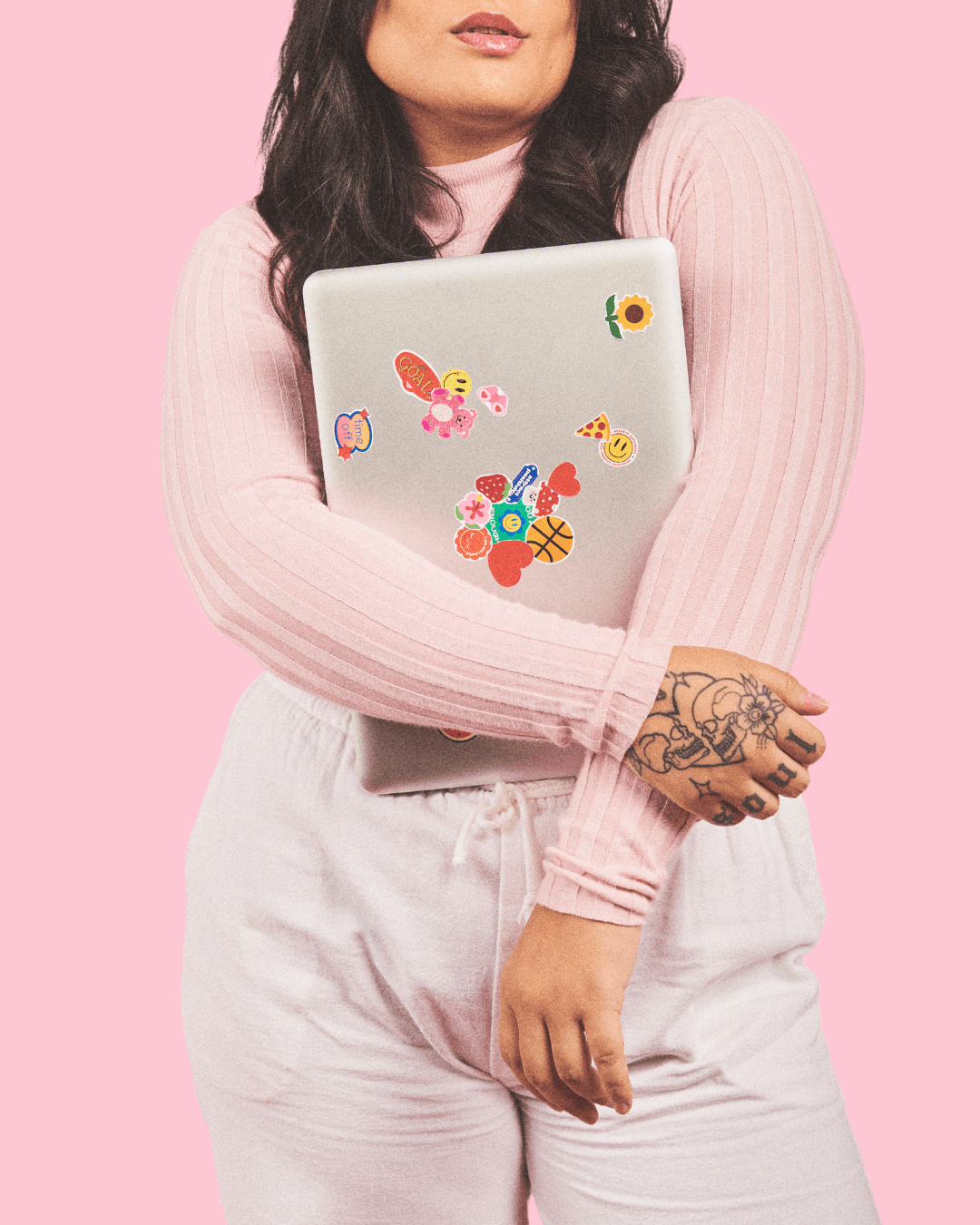 Woman with brown hair and light pink sweater, hugging laptop with stickers all over it.